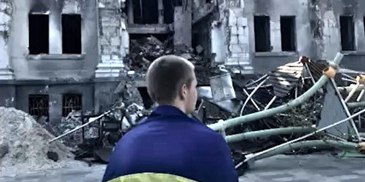 Протест: млад Украинец, завиткан во знамето на Украина, стои пред урнатините на театарот во Мариупол кој беше бомбардиран на 16 март 2022 година. Објавено во видео обраќањето на Претседателот на Украина, Володимир Зеленски, на 18 јули 2022 година.