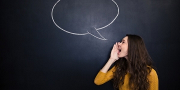 Profile of charming young woman standing and screaming against background of chalkboard with drawn blank speech bubble